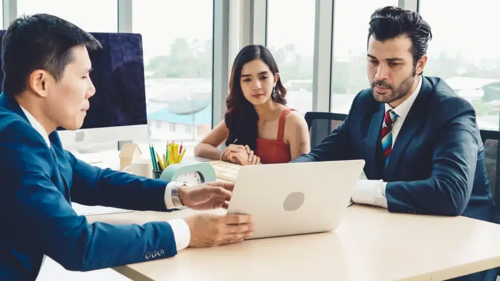 Photo of three business people working at a conference desk as a man shows them his laptop screen
