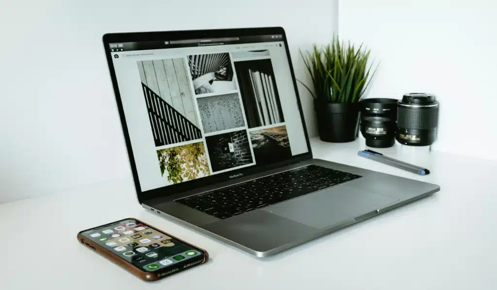 Photo of a MacBook pro with photos open on screen next to an iPhone on a white table with pens a plant and camera