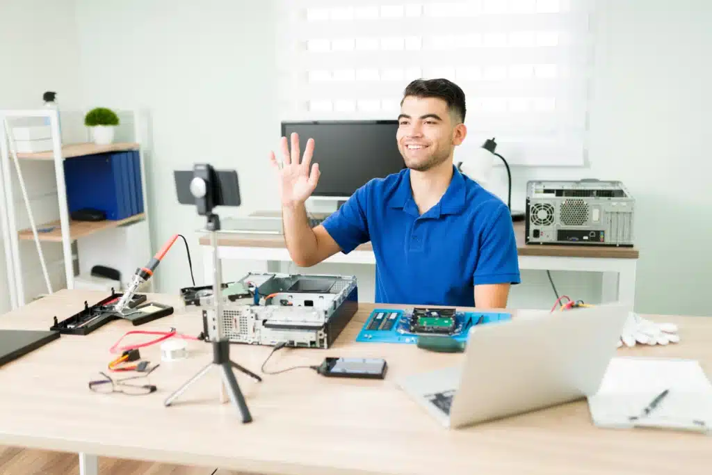 Photo of a guy in a blue shirt waving at an iPhone on a tripod on a desk with a laptop and several computer components