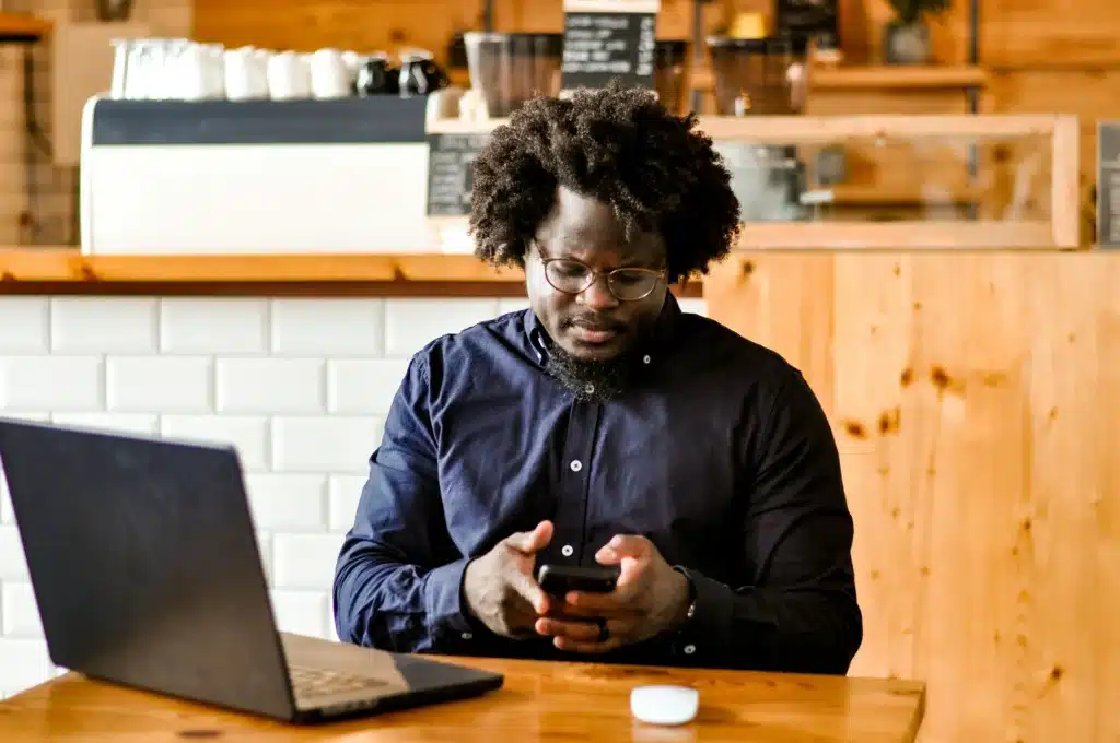 Photo of a man dressed in a dress shirt sitting in a coffee shop with a laptop while typing on a smart phone