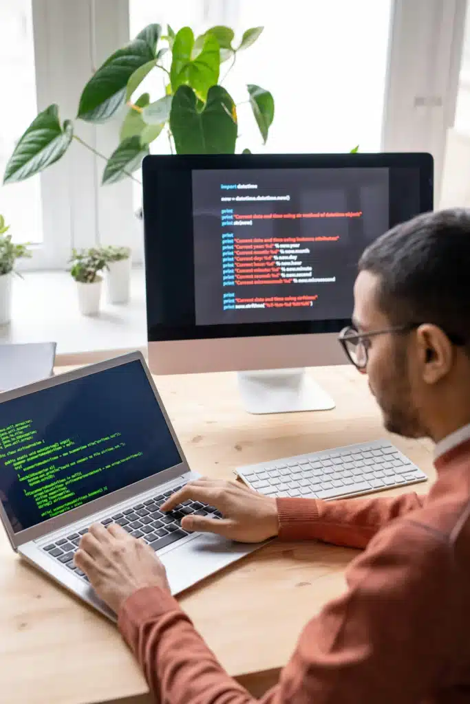 Photo of a man typing code on a MacBook next to an iMac at a desk