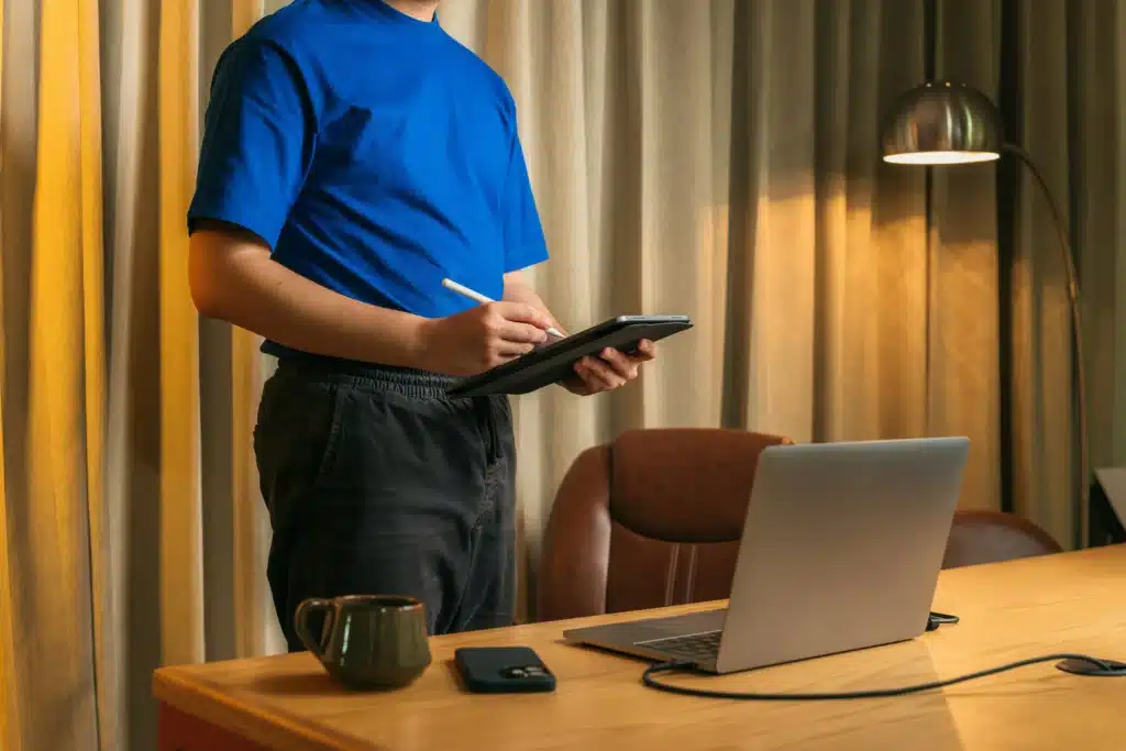 Man in blue t-shirt writing on iPad while standing behind desk with MacBook