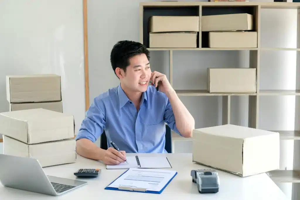 Photo of a man smiling while talking on the phone and writing on a notepad at a desk with a laptop, calculator, boxes, and a card machine