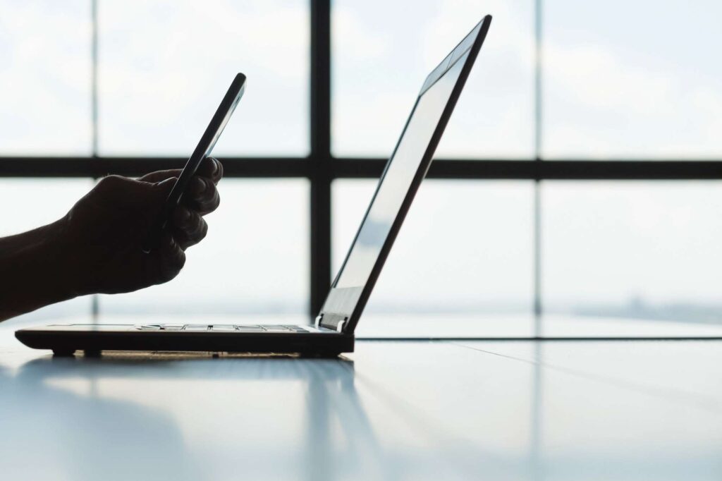 Person using a smartphone beside a laptop, representing the shift from SMS authentication to more secure authenticator apps for improved account security